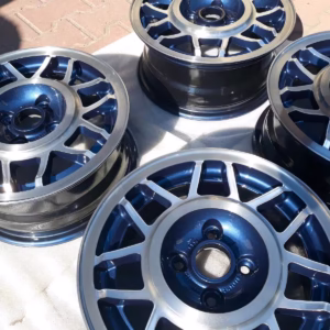 Vier Avus Snowflake Alufelgen Lk5t Inka Blue Golf 171601025H sind im Freien auf einer hellen Fläche angeordnet, wobei das Sonnenlicht ihre reflektierenden blauen und silbernen Oberflächen hervorhebt.