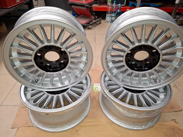 Vier silberne Bmw E12-E3-E9-E28 Felgen Aufbereitet 6x14 Et22 mit Mehrfachspeichen sind in einer Werkstatt paarweise aufrecht auf Karton gestapelt, um ihr frisch aufgearbeitetes Finish zu betonen.