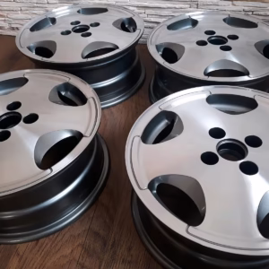 Vier silberne Felgen Fire and Ice Estoril Felgen Golf 1/Golf 2 6x15 sind auf einem Holzboden ausgestellt, mit einer mit Steinmustern versehenen Wand im Hintergrund.