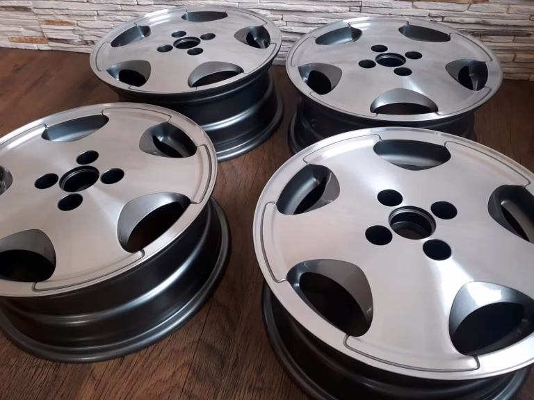 Vier silberne Felgen Fire and Ice Estoril Felgen Golf 1/Golf 2 6x15 sind auf einem Holzboden ausgestellt, mit einer mit Steinmustern versehenen Wand im Hintergrund.