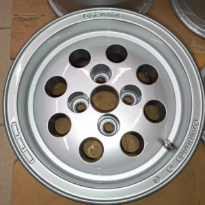 Das Ford Capri Escort Felgen Käseloch 7x13 Et5 ist eine silberne Leichtmetallfelge mit acht kreisrunden Löchern und eingraviertem Branding, ideal für Oldtimer-Liebhaber und auf einer Pappfläche ausgestellt.