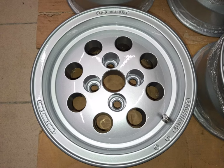 Das Ford Capri Escort Felgen Käseloch 7x13 Et5 ist eine silberne Leichtmetallfelge mit acht kreisrunden Löchern und eingraviertem Branding, ideal für Oldtimer-Liebhaber und auf einer Pappfläche ausgestellt.