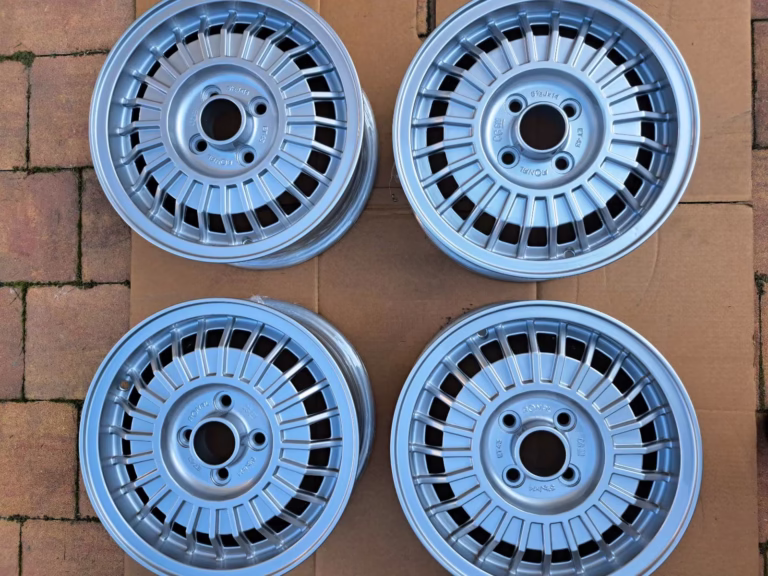 Vier GM Ronal Opel Rekord Felgen Aufbereitet 5.5x14 silberne Leichtmetallräder mit Mehrfachspeichen sind in einem Quadrat auf einer Kartonfläche über Ziegelpflaster angeordnet.