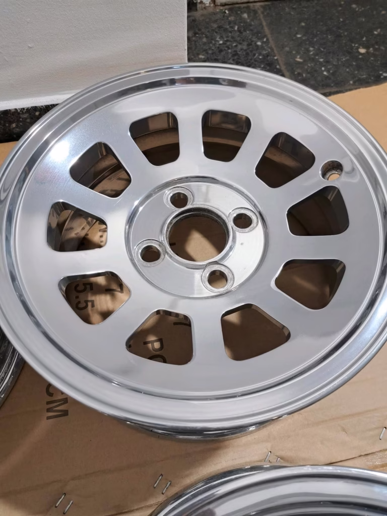 Ein polierter Mini Cooper Alufelgen Hochglanzverdichtet 6x13 verfügt über zehn ovale Speichen und eine zentrale Nabe, die auf einer Kartonoberfläche abgebildet sind - perfekt, um den Stil Ihres Fahrzeugs aufzuwerten.