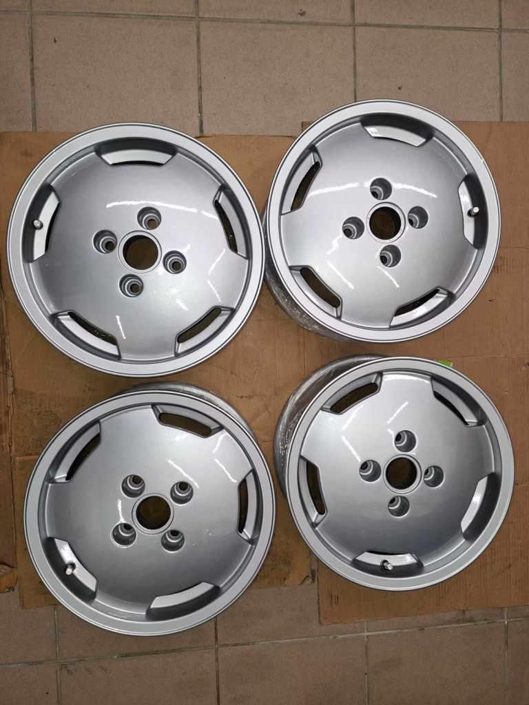 Vier silberne Ronal Audi 80 90 Felgen 393601025A mit je fünf Radmuttern sind in einem rechteckigen Muster auf einem gekachelten Boden mit Kartonunterlage angeordnet - perfekt für Audi-Liebhaber.