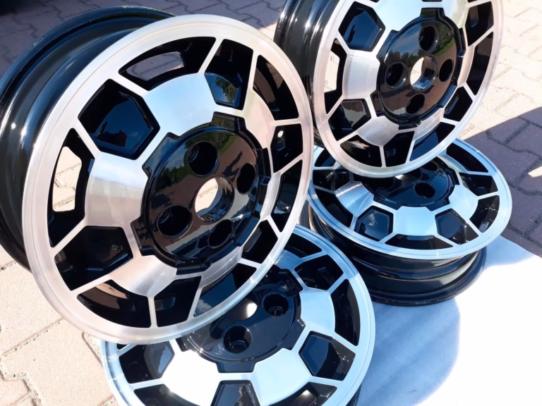 Vier Saab 900 90 Alufelgen Fußbal Felgen 5.5x15, in schwarz und silber, gestapelt auf einer gepflasterten Fläche bei Tageslicht.